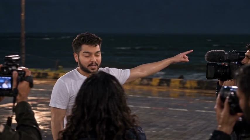 Deep, in a white T-shirt, points out to a dark sea as photographers and a TV camera crowd around him on a wet seafront at night.
